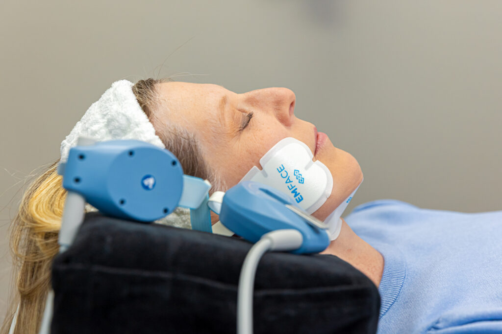 Woman in her 30s or early 40s, lies on a comfortable treatment table. Our provider has attached special applicators to her cheek and submentum for Emface near Arboretum area.