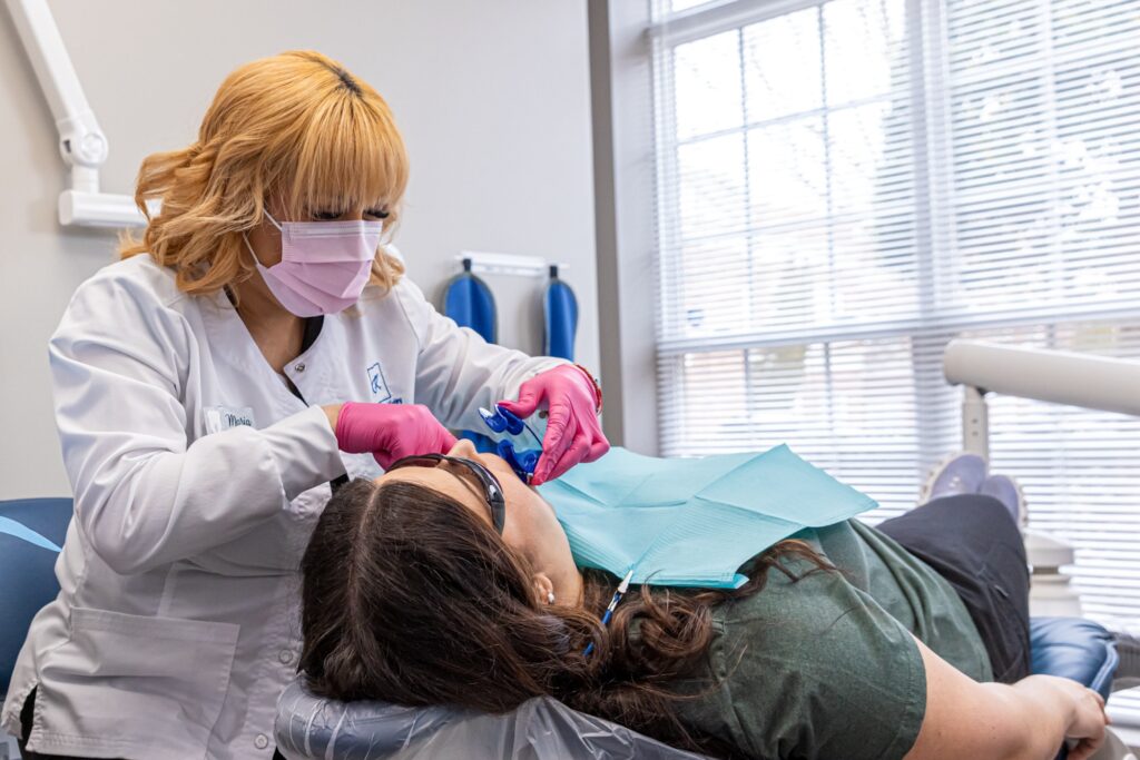 A family dentist near Charlotte is performing a dental procedure for a patient. The patient, relaxing on her back in the dental chair, wears a dental bib as her dentist gently works on her teeth.